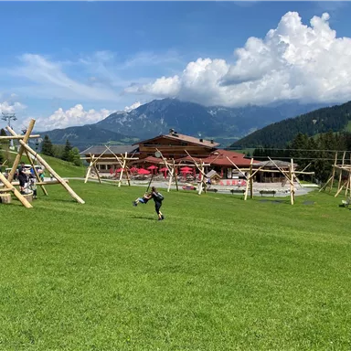 Ein Spielplatz auf einer grünen Wiese mit Holzgerüsten und einem schönen Bergpanorama im Hintergrund. Der Himmel ist blau mit einigen Wolken.