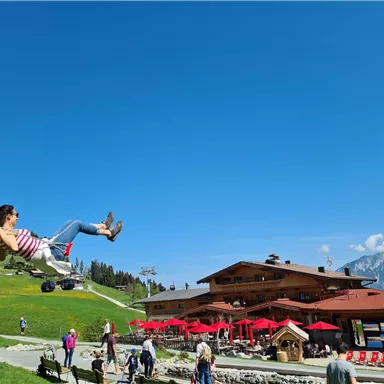 Eine Frau schwingt auf einer Schaukel vor einem Bergrestaurant. Im Hintergrund sind grüne Wiesen und blauer Himmel zu sehen.