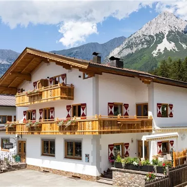 A traditional farmhouse with wooden decorations and colorful shutters. In the background, majestic mountains rise under a blue sky.