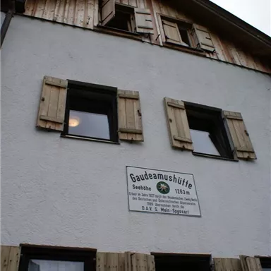 Ein traditionelles Haus mit Holzfensterläden und einem Schild mit dem Namen "Gaudeamushütte". Oben sind kleine Fenster und eine Holzverkleidung zu sehen.