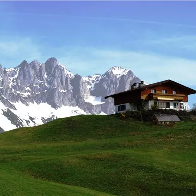 A picturesque house on a hill, surrounded by lush greenery. In the background, snow-capped mountains rise under a clear blue sky.