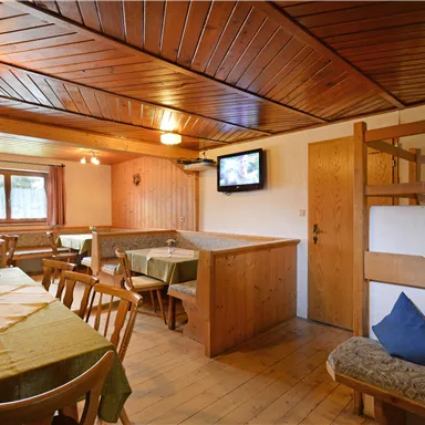 Ein gemütlicher Innenraum mit Holzmöbeln und einer warmen Atmosphäre. Es gibt einen Fernseher an der Wand und Tische mit Stühlen, die zur Geselligkeit einladen.