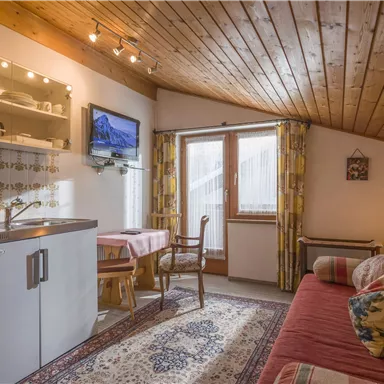 A cozy living environment with a wooden ceiling, a small kitchen, and a television. A sofa and a table create an inviting atmosphere.