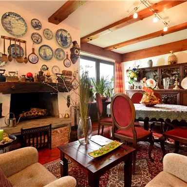 Ein gemütlicher Wohnraum mit einem Tisch und Stühlen. An der Wand hängen dekorative Teller und es gibt eine rustikale Kaminverkleidung.