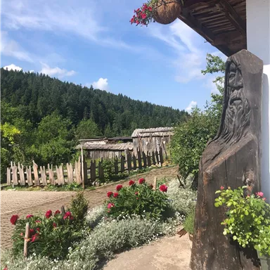 Eine Holzskulptur steht neben einem Blumenbeet mit roten Blumen. Im Hintergrund sieht man eine grüne Landschaft und ein altes Zaun.