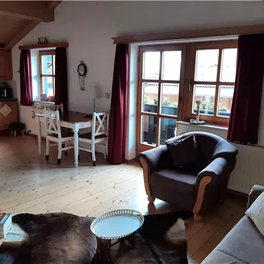 Ein gemütliches Wohnzimmer mit einem Ohrensessel und einem Tisch. Große Fenster sorgen für viel Tageslicht und eine einladende Atmosphäre.