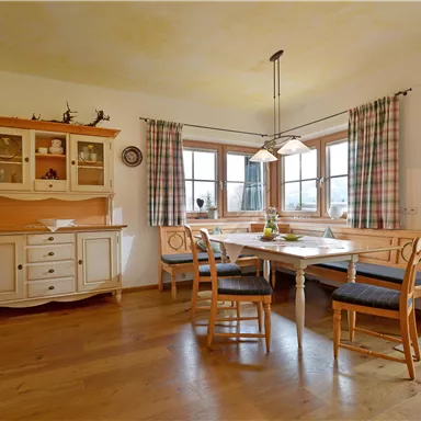 Ein helles Esszimmer mit Holzboden und großen Fenstern. Es gibt einen Tisch mit Stühlen und eine Vitrine im Hintergrund.