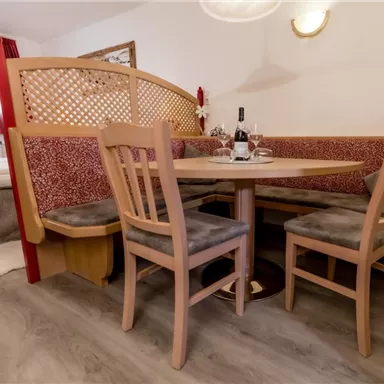 Ein gemütlicher Essbereich mit einem runden Tisch und Holzstühlen. Im Hintergrund sind weiche Sitzpolster und eine Flasche Wein auf dem Tisch zu sehen.