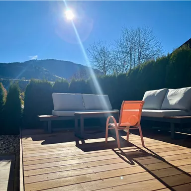 A sunny outdoor area with a wooden terrace. On the terrace, there are two cozy sofas and an orange chair.