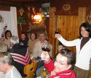 Eine gemütliche Versammlung von Menschen in einem Holzraum, die mit Gläsern anstoßen. Einige Personen lächeln und heben ihre Getränke in die Luft.