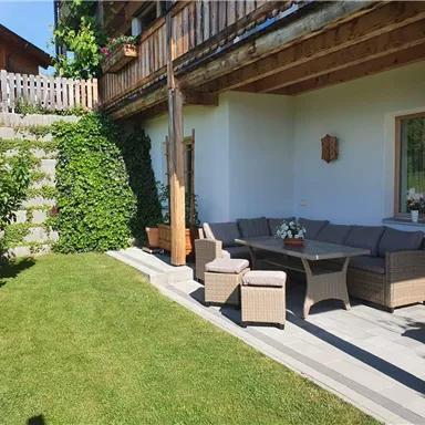 A cozy terrace with a gray seating area and a table. The garden is green with shrubs and a staircase in the background.