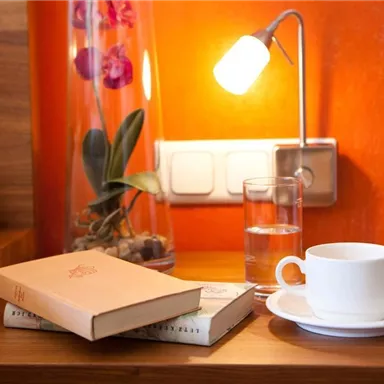 Ein stilvoller Nachttisch mit einem Buch, einer Tasse Kaffee und einem Glas Wasser. Im Hintergrund steht eine Orchidee in einer Vase und eine kleine Lampe leuchtet.