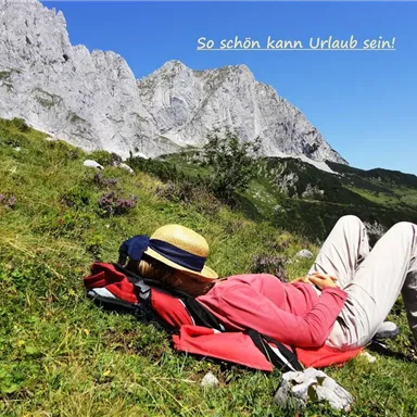 Eine Person liegt entspannt auf einer Wiese und genießt die Aussicht auf die Berge. Der Himmel ist blau und die Umgebung ist grün und bergig.