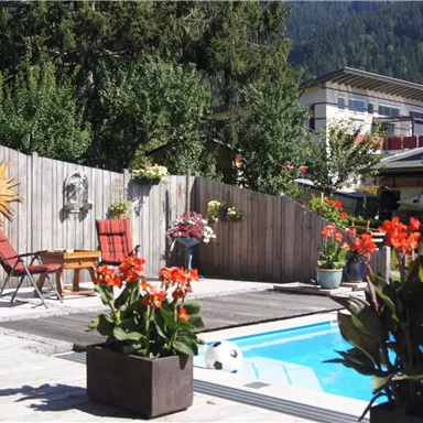 An inviting pool area with blooming plants and cozy chairs. In the background, there are trees and a building.