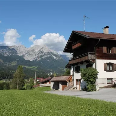 An idyllic house in the mountains with green meadows and majestic peaks in the background. The sky is clear and sunny.