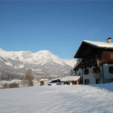 A picturesque winter landscape with snow-covered mountains in the background. In the foreground stands a traditional wooden house.