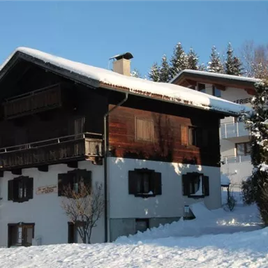 A traditional chalet in the snow, surrounded by trees. The sky is clear and the sun shines through the winter landscape.