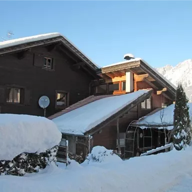 A traditional chalet in the snow with a clear blue sky. Snow-covered mountains can be seen in the background.