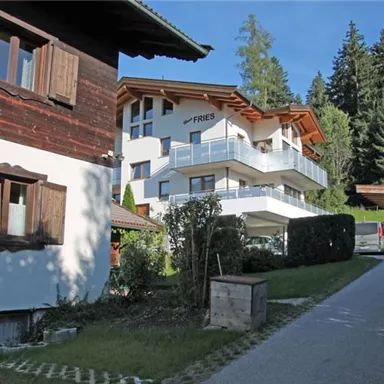 A modern building with a balcony, surrounded by trees. In the foreground stands a traditional wooden house and a cozy street.