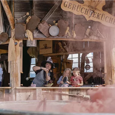 Eine rustikale Hütte mit dem Schild „Gerücheküche“ und mehreren Kindern, die neugierig zuschauen. Eine Frau bereitet etwas vor, während sich bunte Dämpfe um sie herum bilden.