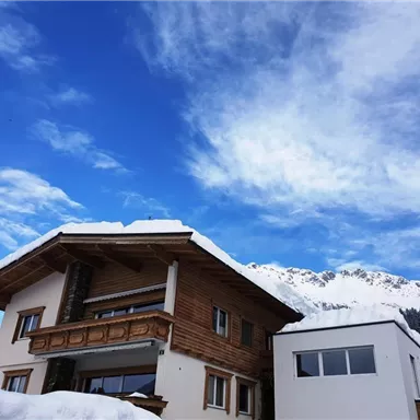 A cozy house in the snow with a traditional wooden design. The sky is clear and blue, while the mountains are visible in the background.