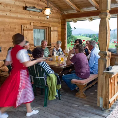 Eine Gruppe von Menschen sitzt an einem Holztisch auf einer Veranda und genießt gemeinsam eine Mahlzeit. Im Hintergrund sind grüne Felder und Blumen zu sehen.