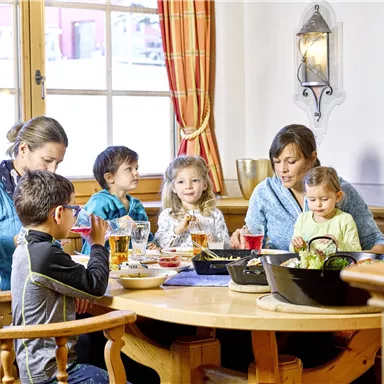 Eine fröhliche Gruppe von Erwachsenen und Kindern sitzt um einen Tisch und genießt eine Mahlzeit zusammen. Das Ambiente ist gemütlich und einladend.