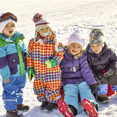 Vier fröhliche Kinder spielen im Schnee, angezogen in bunte Winterkleidung. Sie sitzen auf Schlitten und genießen den sonnigen Tag.