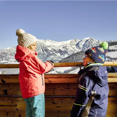 Zwei Kinder stehen an einer Holzbarriere und schauen auf eine schneebedeckte Berglandschaft. Sie tragen warme Kleidung und Mützen, während der klare blaue Himmel über ihnen leuchtet.