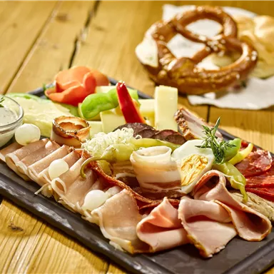 Eine abwechslungsreiche Platte mit Wurst, Käse und frischem Gemüse. Im Hintergrund sind ein Brezel und ein Brötchen zu sehen.