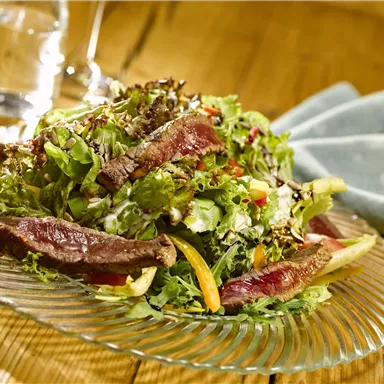 Ein frischer Salat mit saftigem Fleisch und buntem Gemüse. Er liegt auf einem eleganten Glas-Teller und wird von einem schlichten Tischtuch begleitet.