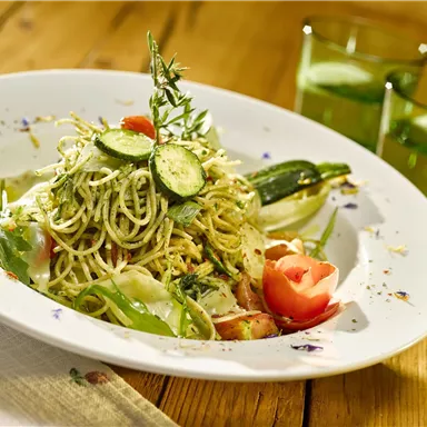 Ein leckerer Teller Pasta mit frischem Gemüse und Kräutern. Im Hintergrund stehen zwei grüne Getränke.