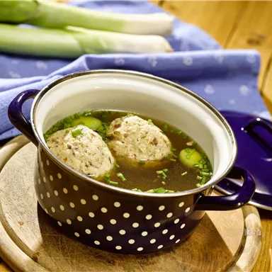 Eine Schüssel mit Brühe und zwei Klößen darin. Im Hintergrund sind frische Lauchzwiebeln zu sehen.