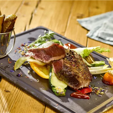 Ein Teller mit zartem Fleisch, buntem Gemüse und einer Beilage von Ofenkartoffeln. Das Gericht ist auf einem schwarzen Tablett angerichtet und sieht appetitlich aus.