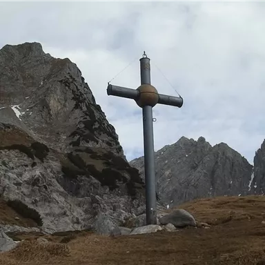 A large cross stands on a high mountain meadow. In the background, impressive rocks and a cloudy sky can be seen.