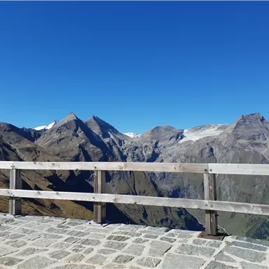 An impressive mountain landscape with high peaks and clear blue sky. In the foreground, a wooden railing and stone floor.