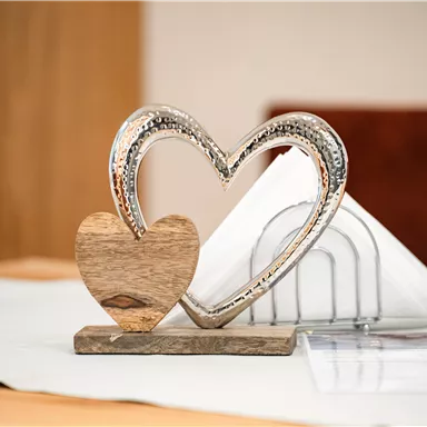 A decorative heart sculpture made of wood and metal is standing on a table. In the background, folded napkins and a neutral table surface can be seen.