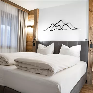 A modern bedroom with a large bed and two pillows. A stylized mountain graphic hangs on the wall, and the furniture is made of wood.