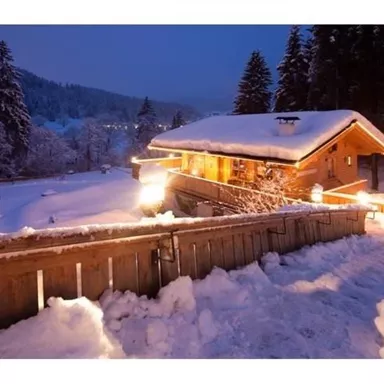 A cozy wooden cottage in the snowy landscape at night. The surroundings are quiet and covered with fresh snow, while warm lights illuminate the house.