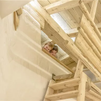 Zwei Kinder schauen neugierig von oben durch eine Holztreppe. Der helle Raum hat eine warme Atmosphäre durch die Holzelemente.