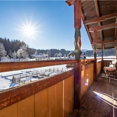 Ein Balkon mit Holzgeländer, der einen Blick auf eine verschneite Landschaft bietet. Die Sonne strahlt klar am Himmel und sorgt für eine helle, einladende Atmosphäre.