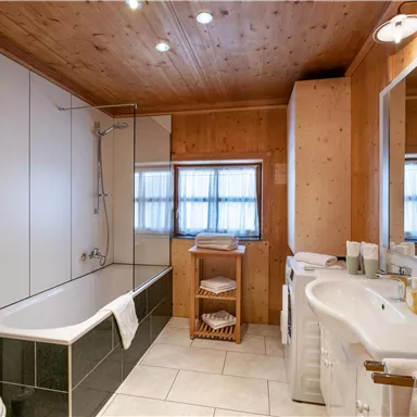Ein modernes Badezimmer mit einer Badewanne und einem Waschbecken. Die Wände sind hell mit Holzverkleidung und großen Fenstern für natürliches Licht.
