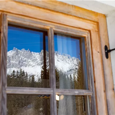 Ein Fenster mit einem Holzrahmen, das die verschneiten Berge und den klaren blauen Himmel spiegelt. Im Hintergrund sind auch Wälder sichtbar.