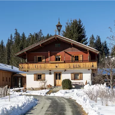 Ein gemütliches Holzhaus mit roter Fassade und einem Balkon. Es liegt in einer schneebedeckten Landschaft, umgeben von Bäumen.