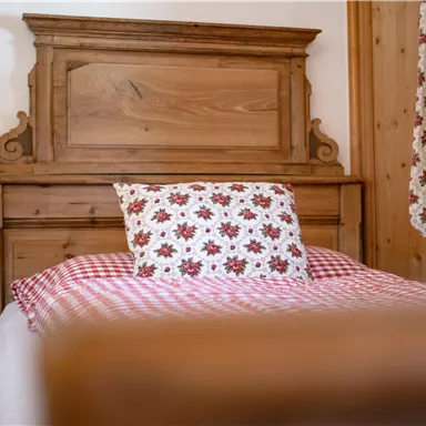 Ein gemütliches Schlafzimmer mit einem Holzkopfteil und einem karierten Bettlaken. Das Kissen hat ein blühendes Muster und eine warme, einladende Atmosphäre.