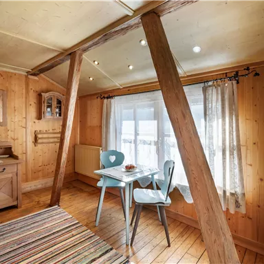 Ein gemütlicher Raum mit holzverkleideten Wänden und Fußboden. Ein kleiner Tisch mit zwei Stühlen steht vor einem Fenster, das viel Tageslicht hereinlässt.