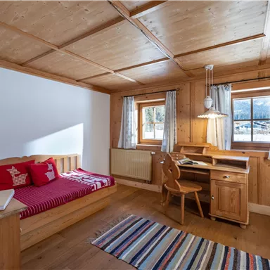 Ein gemütliches Schlafzimmer aus Holz mit einem Einzelbett, einem Schreibtisch und einem Stuhl. Große Fenster sorgen für viel Tageslicht und einen Blick nach draußen.
