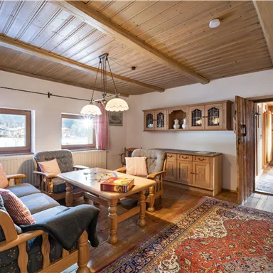 Ein gemütliches Wohnzimmer mit Holzdecken und einem großen Tisch. Die Einrichtung ist rustikal mit bequemen Sesseln und einem bunten Teppich.