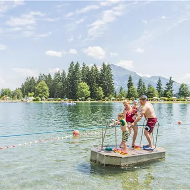 Ein kleiner Steg im Wasser mit zwei Erwachsenen und einem Kind, die Spaß haben. Im Hintergrund sind Bäume und Berge zu sehen, bei schönem Wetter.
