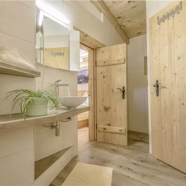 Ein modernes Badezimmer mit Holzdetails und einer großen Spiegelwand. Die Wände sind hell gefliest, und eine Pflanze steht auf dem Waschbecken.